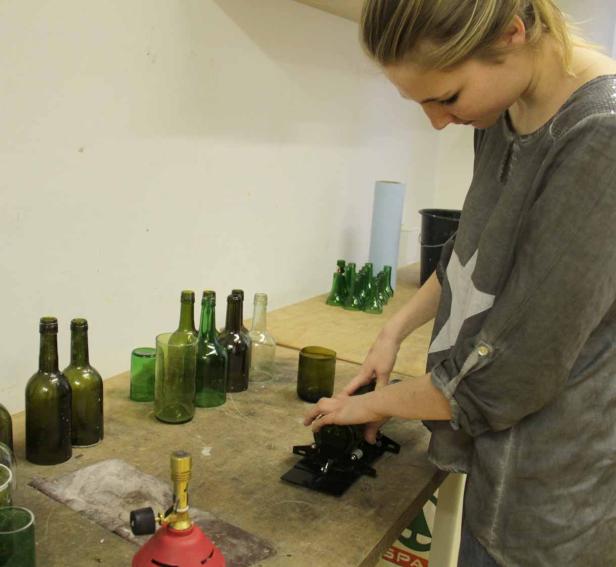 Eine Frau bearbeitet eine grüne Glasflasche mit einem Schneidegerät, um sie in ein Glas zu verwandeln.