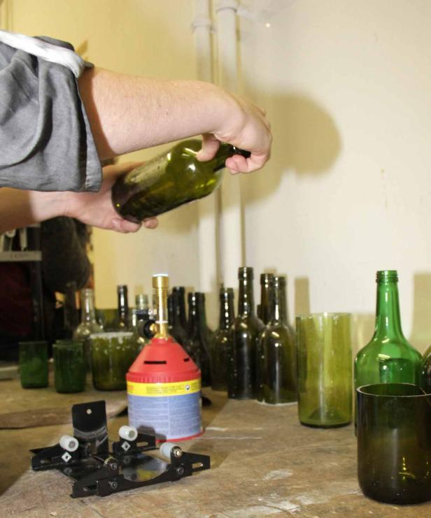 Eine Person bearbeitet eine grüne Glasflasche mit einem Schneidewerkzeug und einem Gasbrenner.