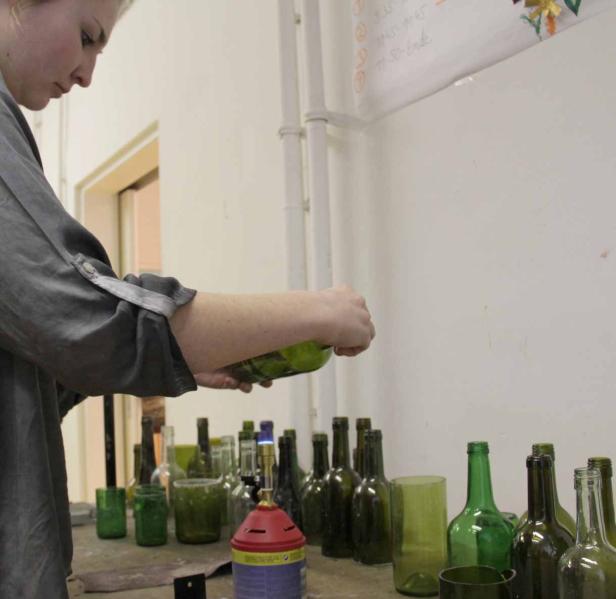 Eine Frau gießt etwas aus einer grünen Flasche über einem Tisch voller Glasflaschen.