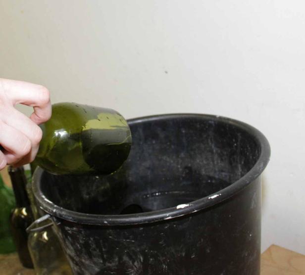 Eine Hand leert eine grüne Glasflasche in einen schwarzen Eimer.