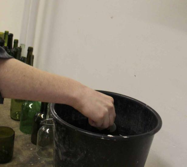 Eine Hand greift in einen schwarzen Eimer, im Hintergrund stehen grüne Flaschen.