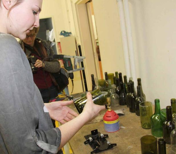 Eine Frau hält eine grüne Glasflasche in der Hand, umgeben von weiteren Flaschen auf einem Tisch.
