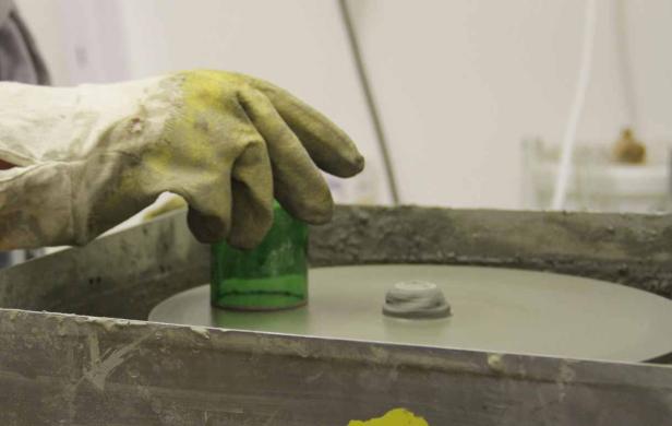 Eine Hand mit Schutzhandschuh poliert einen grünen Gegenstand auf einer Schleifscheibe.