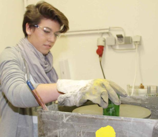 Eine Frau schleift mit Schutzbrille und Handschuhen ein grünes Glasobjekt.