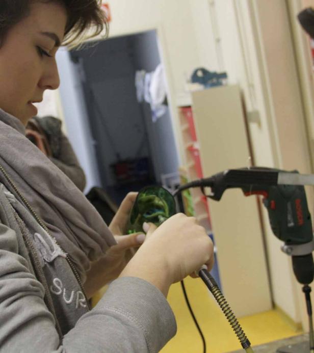 Eine Frau bearbeitet eine grüne Glasmaske mit einem Werkzeug.