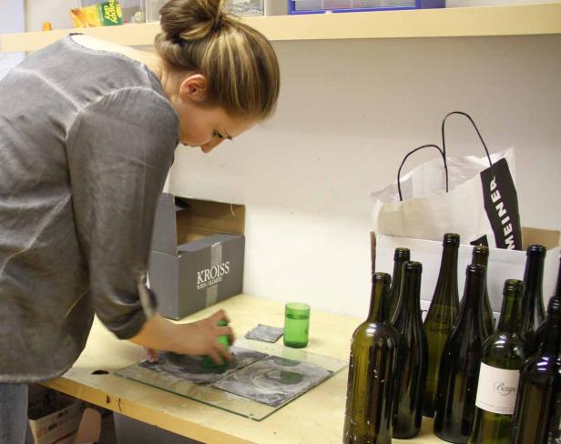 Eine Frau bearbeitet eine Glasplatte mit Farbe, umgeben von Weinflaschen.