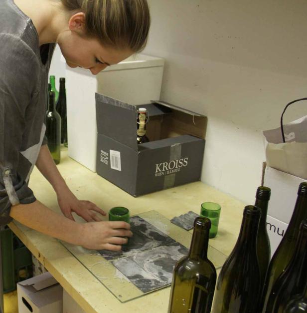 Eine Frau bearbeitet ein grünes Glas auf einer Werkbank, umgeben von Weinflaschen.