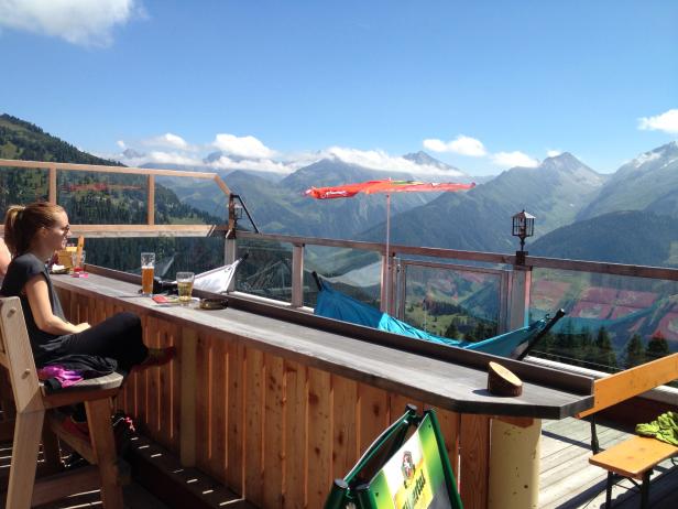 Eine Frau sitzt auf einer Terrasse mit Bergblick und trinkt ein Bier.