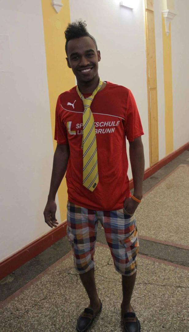 Ein lächelnder junger Mann mit Krawatte, T-Shirt und karierten Shorts steht in einem Flur.