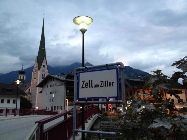 Ein Ortsschild von Zell am Ziller vor einer abendlichen Stadtansicht.