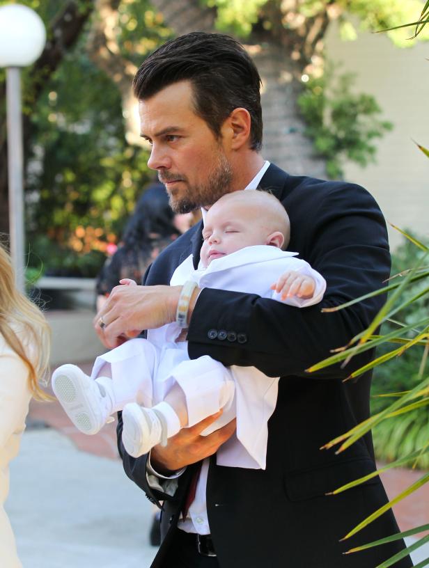 Josh Duhamel trägt ein schlafendes Baby im weißen Taufgewand.