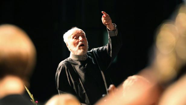 Ein Dirigent mit weißem Bart dirigiert ein Orchester.