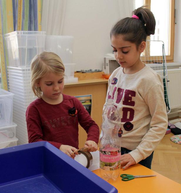 Zwei Mädchen basteln mit einer Plastikflasche, Klebeband und einer Schere an einem Tisch.