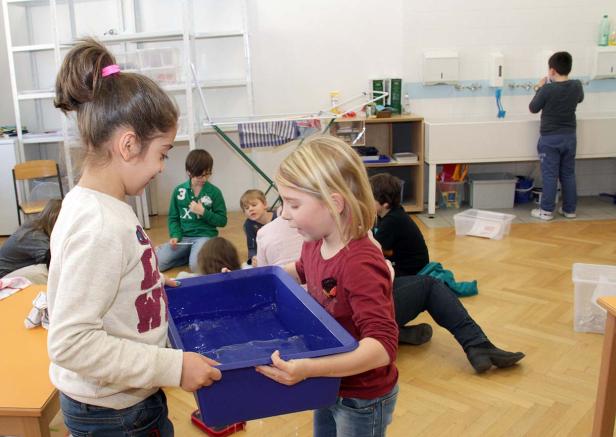 Zwei Mädchen tragen gemeinsam eine blaue Wanne mit Wasser in einem Klassenzimmer.