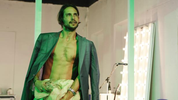 Ein Mann mit offenem Hemd und Sakko steht in einem Raum mit Mikrofon und hält ein Glas in der Hand.