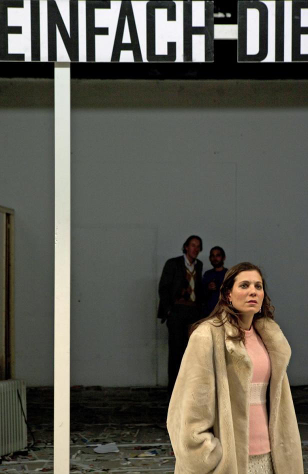 Eine Frau in einem hellen Mantel steht vor einer Wand mit der Aufschrift „Einfach Die“.