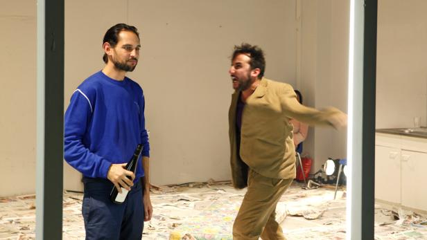 Ein Mann mit einer Flasche Wein steht neben einem schreienden Mann in einem chaotischen Raum.