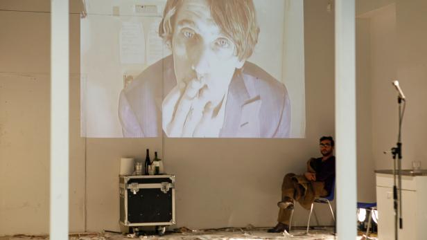 Ein Mann sitzt vor einer Projektion eines Mannes, der in die Kamera schaut.