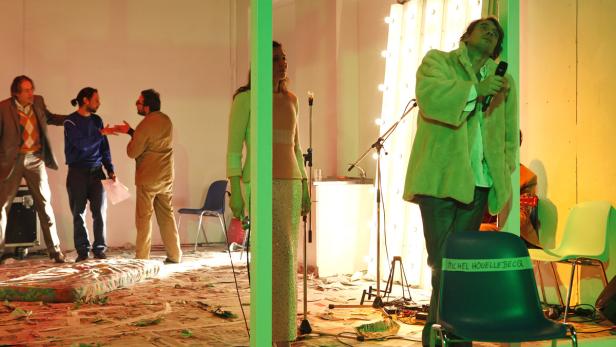 Eine Bühnenszene mit mehreren Personen, darunter ein Sänger mit Mikrofon und ein Stuhl mit der Aufschrift „Michel Houellebecq“.