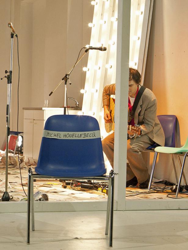 Ein Mann spielt Gitarre auf einer Bühne; ein blauer Hocker mit der Aufschrift „Michel Houellebecq“ steht im Vordergrund.