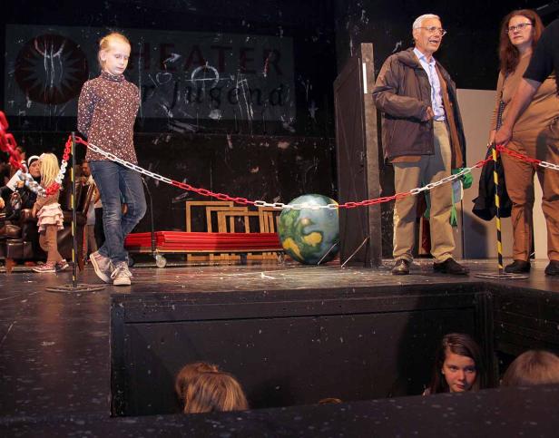 Eine junge Schauspielerin steht auf einer Bühne, während im Hintergrund Zuschauer sitzen.