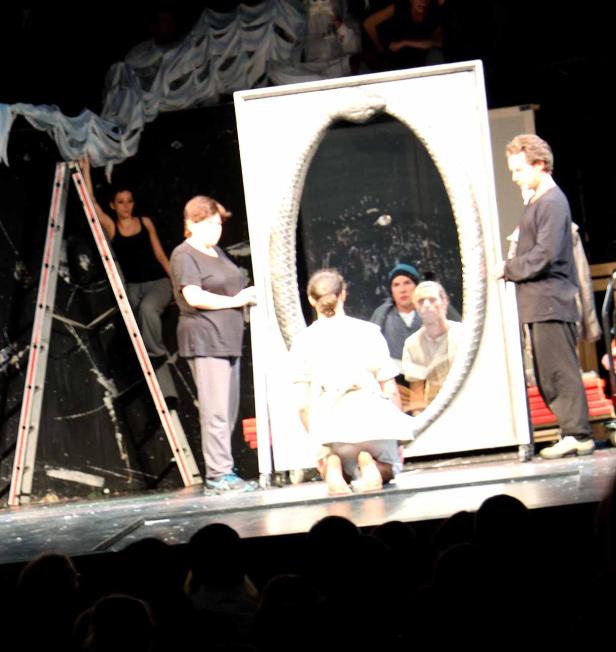Eine Theaterszene mit Schauspielern und einem großen Spiegel auf der Bühne.