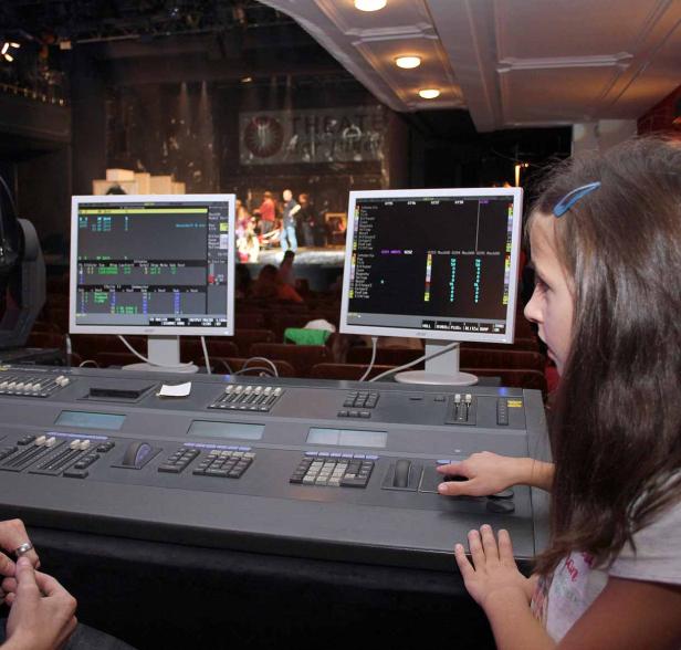 Ein Mädchen bedient ein Mischpult im Theater, während auf der Bühne eine Aufführung stattfindet.