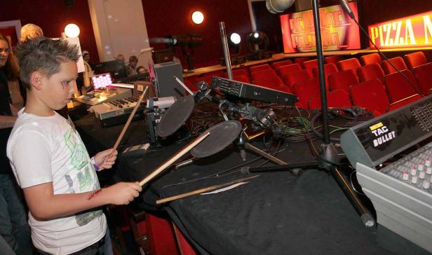 Ein Junge spielt Schlagzeug auf einer Bühne mit rotem Hintergrund.