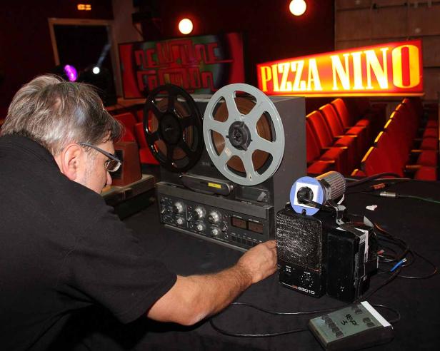 Ein Mann bedient eine Tonbandmaschine vor einem „Pizza Nino“-Schild.