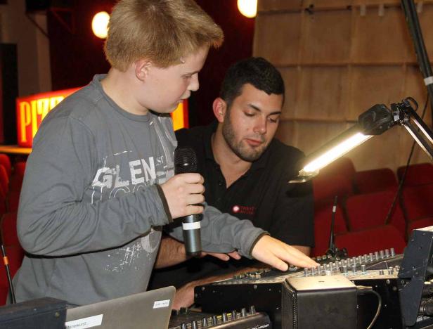 Ein Junge mit Mikrofon bedient ein Mischpult, während ein Mann ihm zusieht.