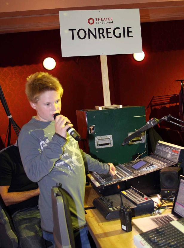 Ein Junge spricht in ein Mikrofon vor einem Mischpult mit dem Schild „Tonregie“.
