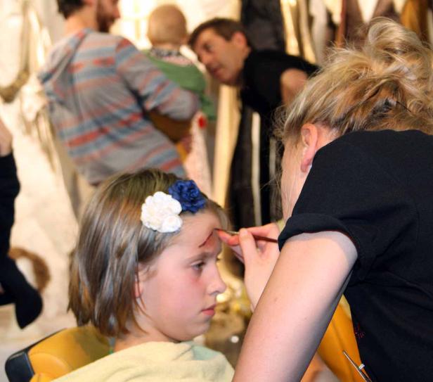 Ein Mädchen mit Blumen im Haar wird geschminkt, um eine Wunde auf der Stirn darzustellen.