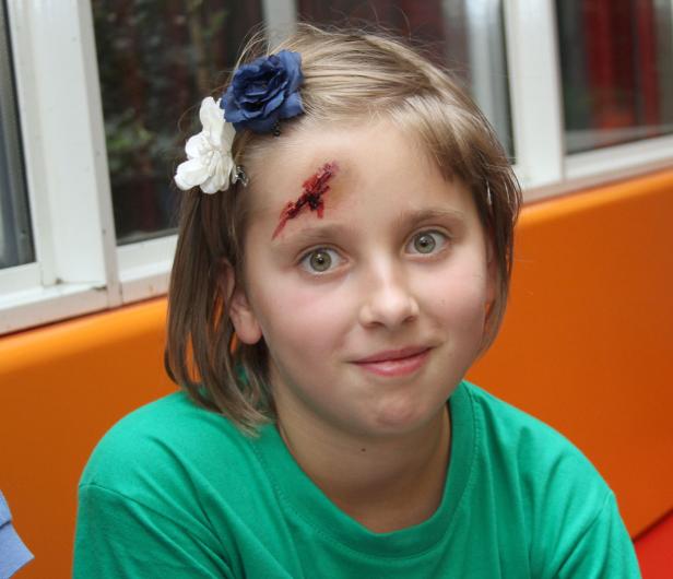 Ein junges Mädchen mit Blumen im Haar und einer Wunde auf der Stirn lächelt.