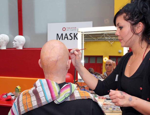 Eine Maskenbildnerin schminkt einen Mann mit Glatze für das „Theater der Jugend“.