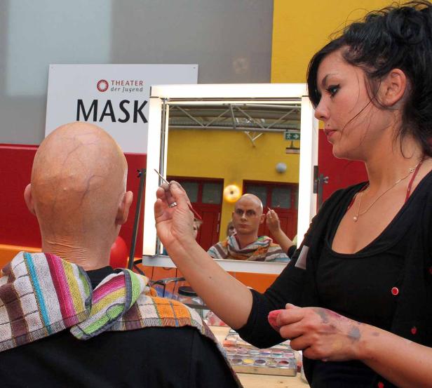 Eine Maskenbildnerin schminkt einen Mann mit Glatze vor einem Spiegel.
