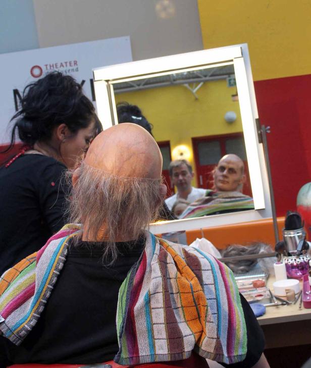Ein Schauspieler mit Glatze und Make-up sitzt vor einem Spiegel in der Maske.