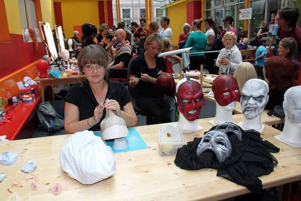 Eine Frau arbeitet an einer Maske, während im Hintergrund weitere Masken und Menschen zu sehen sind.