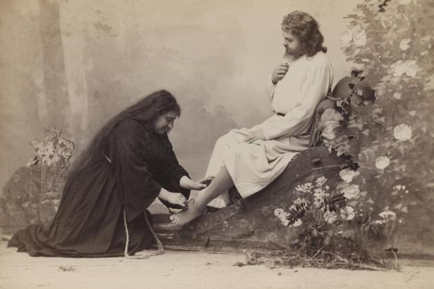 Eine Frau wäscht die Füße einer sitzenden Person vor einem Blumenhintergrund.