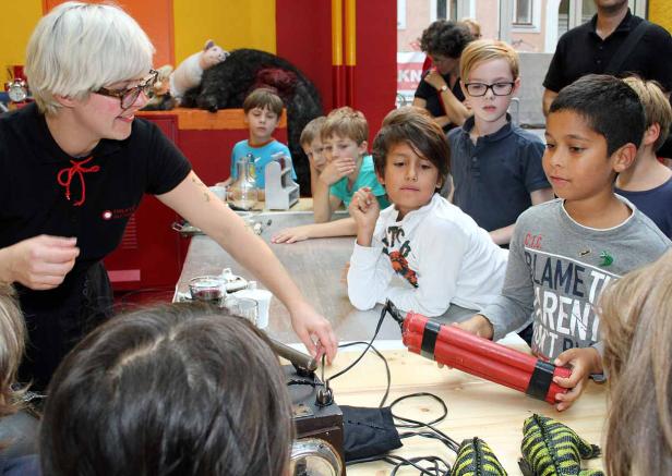 Eine Frau erklärt einer Gruppe von Kindern etwas an einem Tisch mit Requisiten.