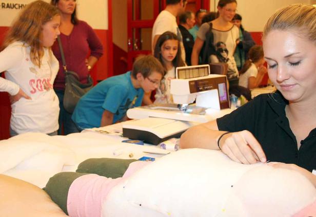 Eine junge Frau näht ein Stofftier, während Kinder eine Nähmaschine beobachten.