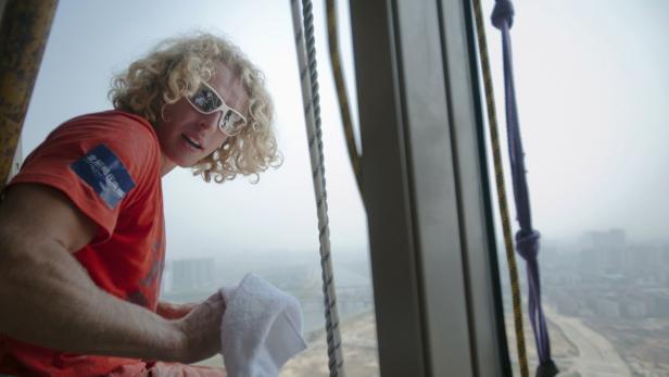 Ein Fensterputzer mit blonden Locken und Sonnenbrille reinigt ein Fenster in großer Höhe.
