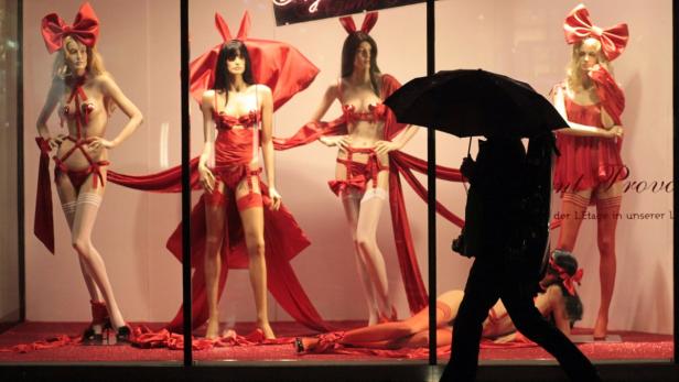 Koffer (1) Ein Schaufenster mit Mannequins in roter Dessous-Mode und großen Schleifen, gesehen durch einen Passanten mit Regenschirm.