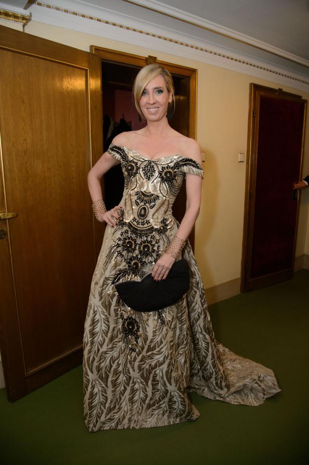 Eine Frau in einem goldenen Abendkleid mit schwarzer Clutch posiert vor zwei Türen.