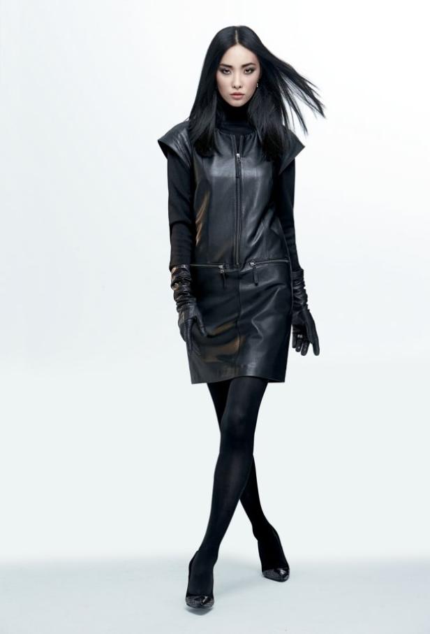 Eine Frau mit dunklen Haaren trägt ein schwarzes Lederkleid, Strumpfhose und Handschuhe.