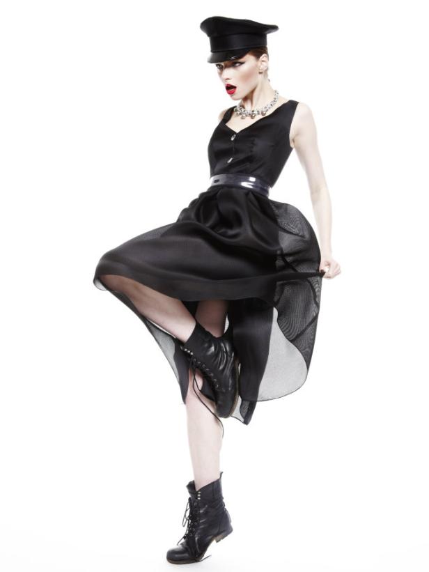 Eine Frau in schwarzem Kleid, Stiefeln und Mütze posiert auf einem Bein.