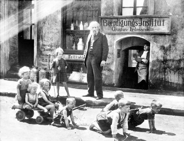 Kinder spielen vor dem „Beerdigungs-Institut Gustav Erdmann“ in einer Straßenszene.