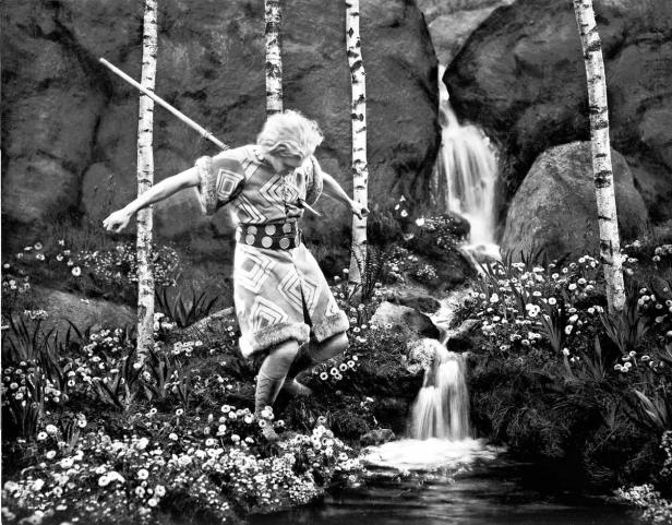 Eine junge Frau in Tracht balanciert an einem Bach mit Wasserfall.