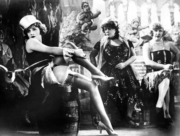 Eine Frau sitzt auf einem Fass, während andere Frauen im Hintergrund stehen.