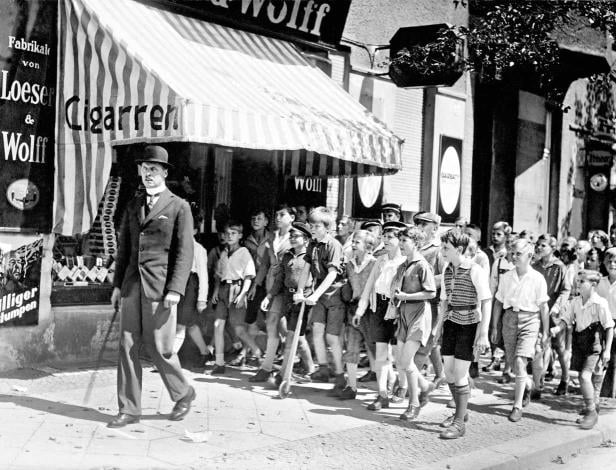 Ein Mann mit Hut geht vor einer Gruppe von Jungen an einem Zigarettengeschäft vorbei.
