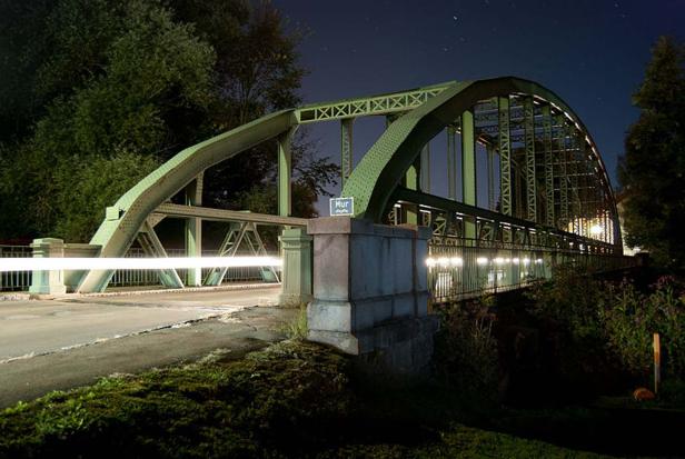 Eine grüne Brücke über die Mur bei Nacht mit Lichtspuren eines vorbeifahrenden Autos.